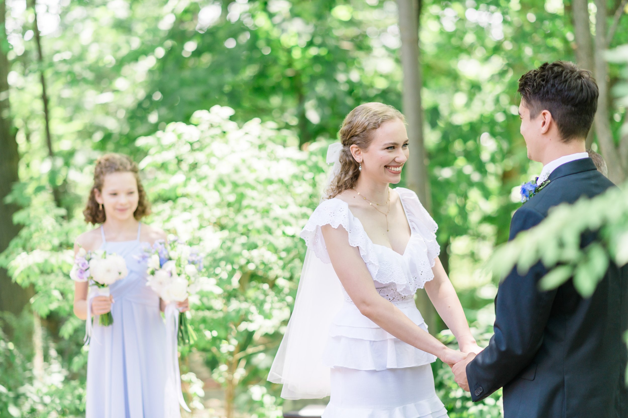 Simone and Josh holding hands during the ceremony, Simone's little sister as an onlooker in the background, holding all the flowers.