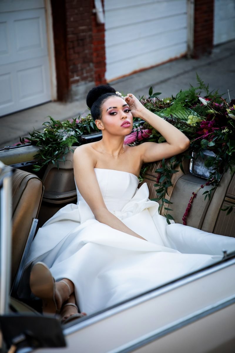 bride reclining in the backset of the vintage car with burgundy, pink and green flowers strewn across the back of the back seat