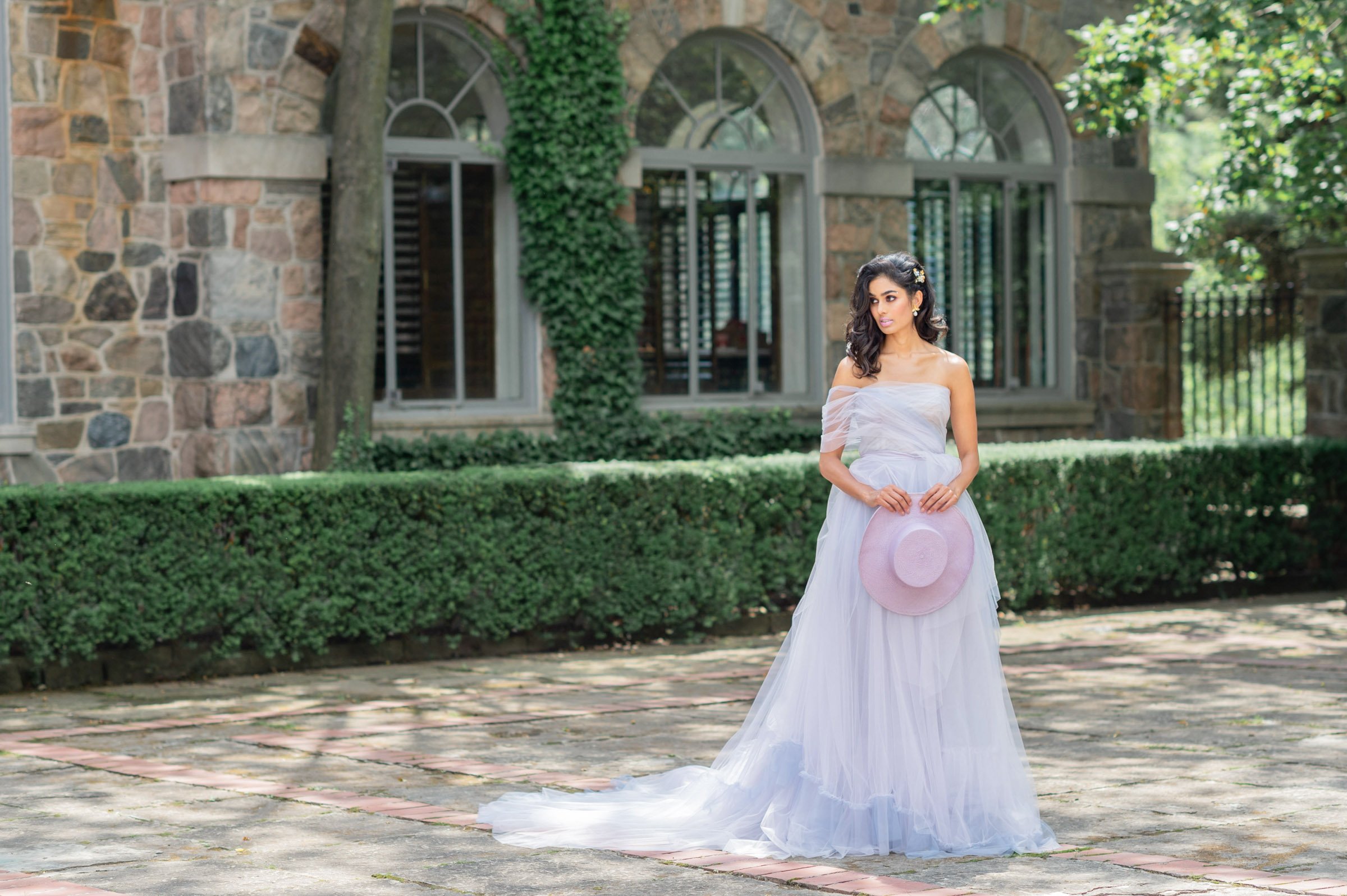 brunette model seen in lilac dress holding a straw hat in front garden of Graydon hall manor.