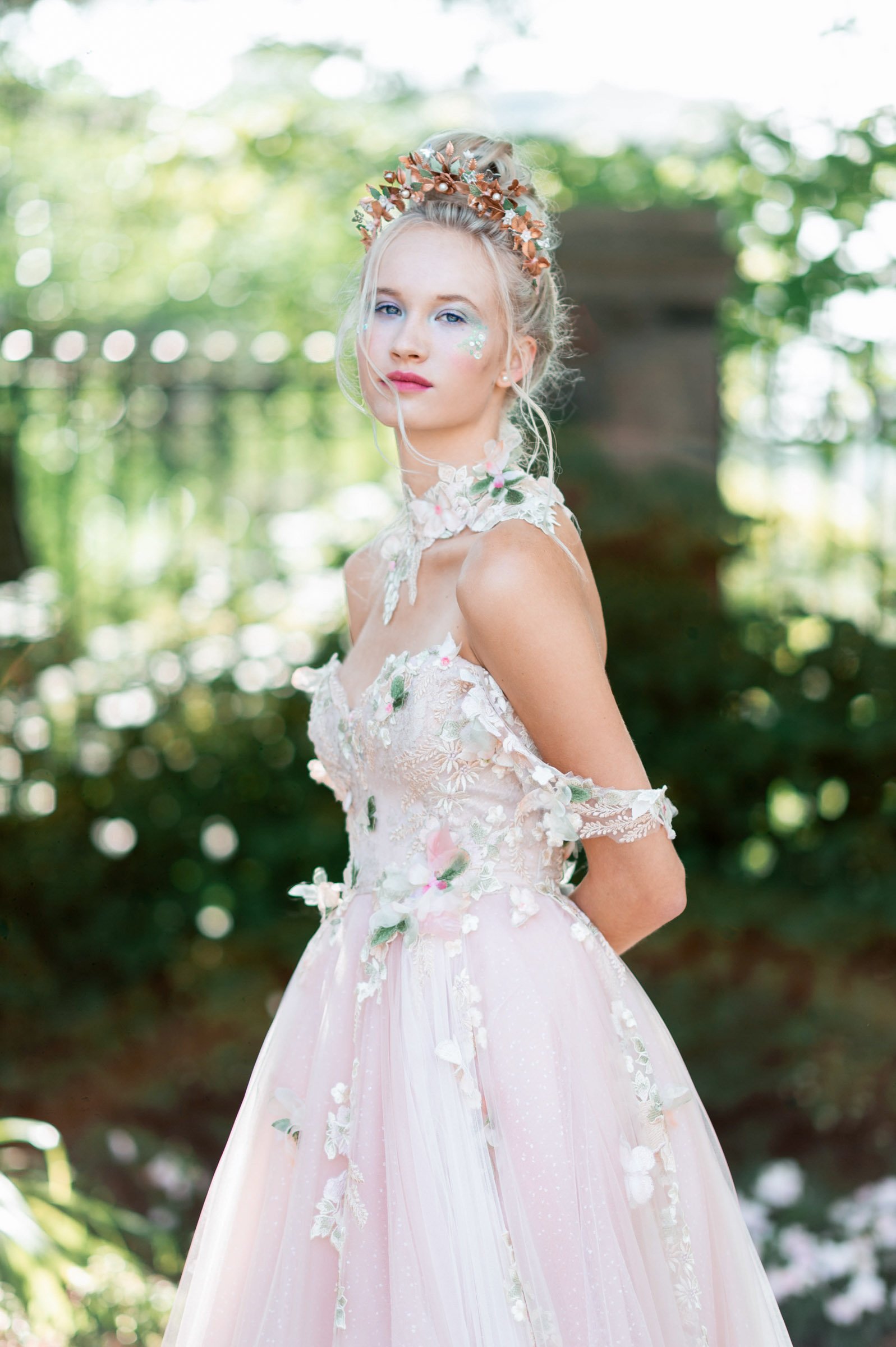 blonde model seen with tiara with leaves and and shimmery eye makeup and hilighter in palest mint green and berry stained lip, wearing a pale pink gown with green leaf and floral lace appliquee with a lace collar with more appliquee.