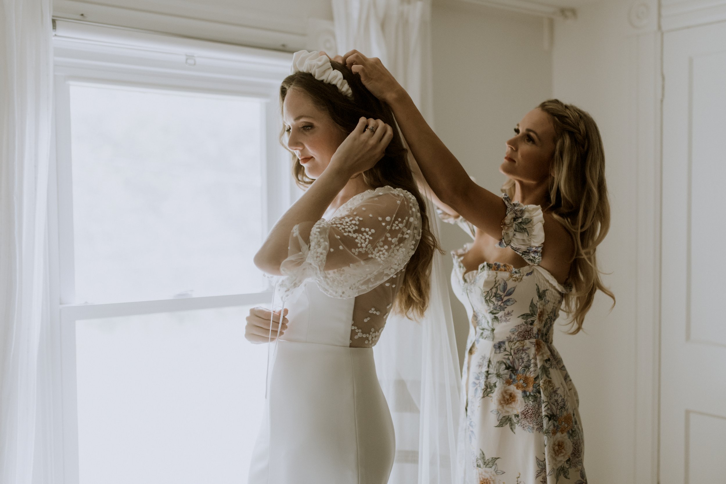 the bride and her sister, helping her with the finishing touches- of veil and velvet hair band.