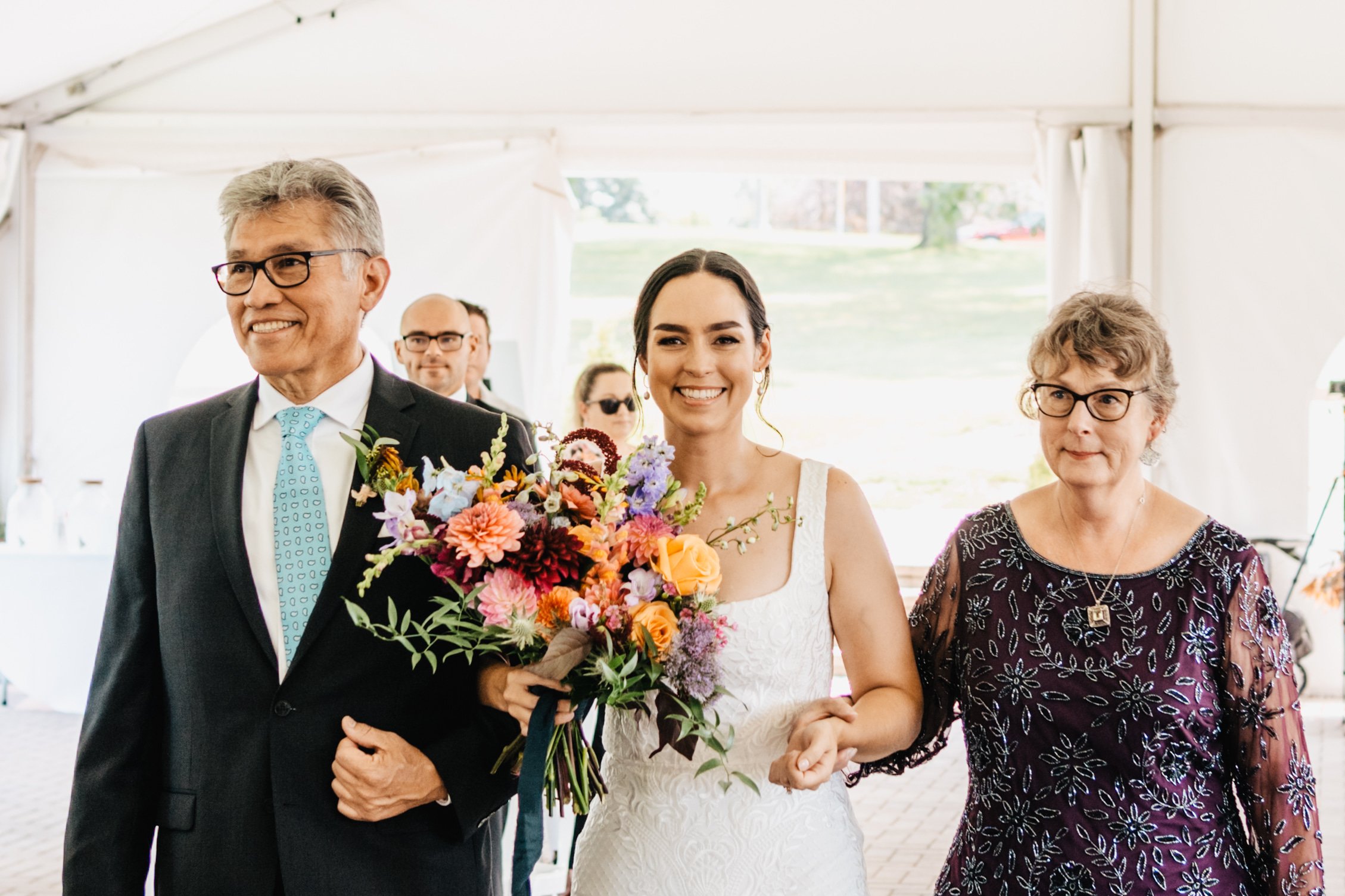 mother and father of the bride walk her down the aisle. Bride holds a beautiful colourful bouquet of flowers in one hand and her mother's hand in the other.