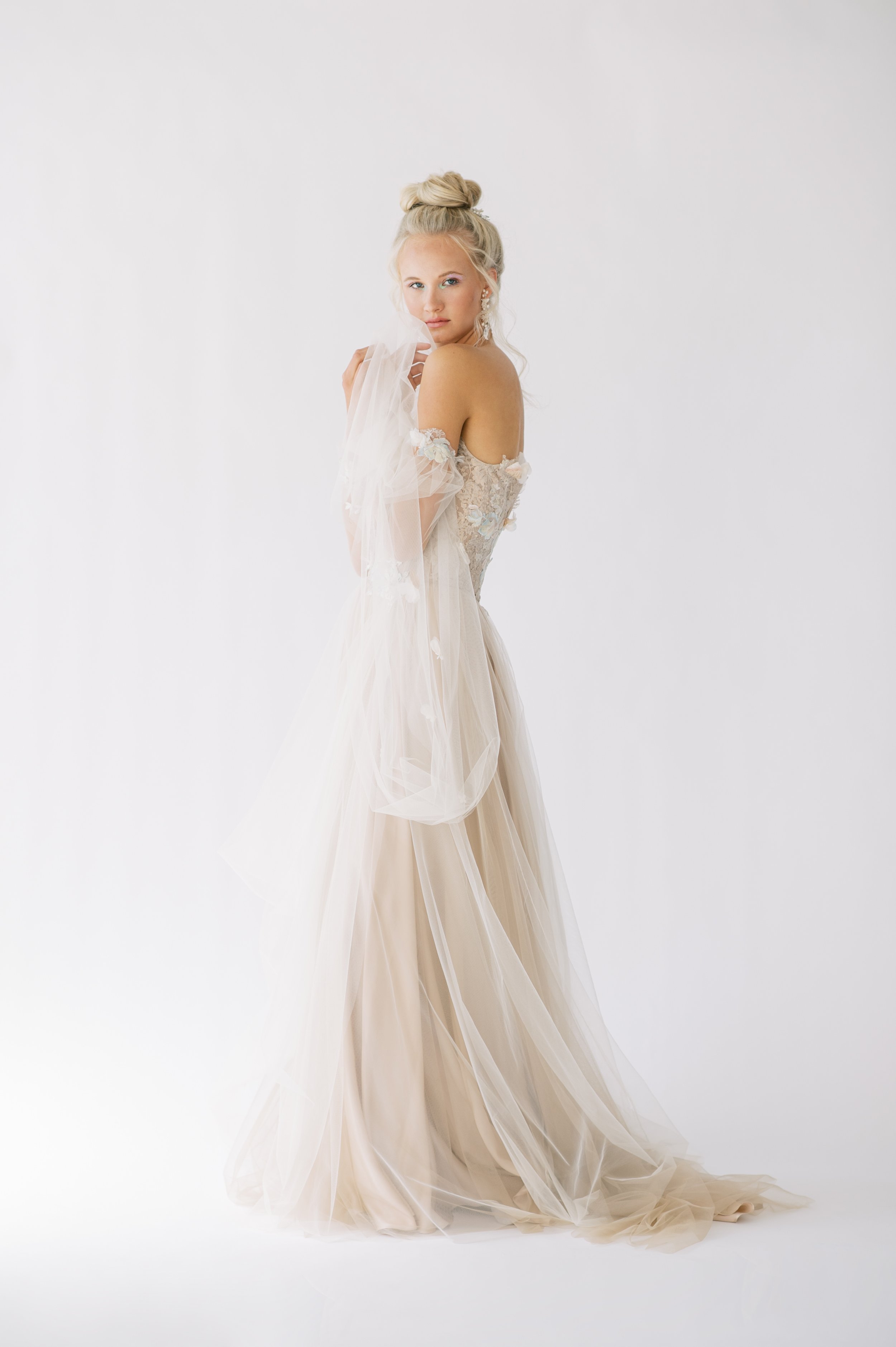 blonde model with high bun and long flowery earrings in a long, off the shoulders, beige and pale blue lace and tulle gown