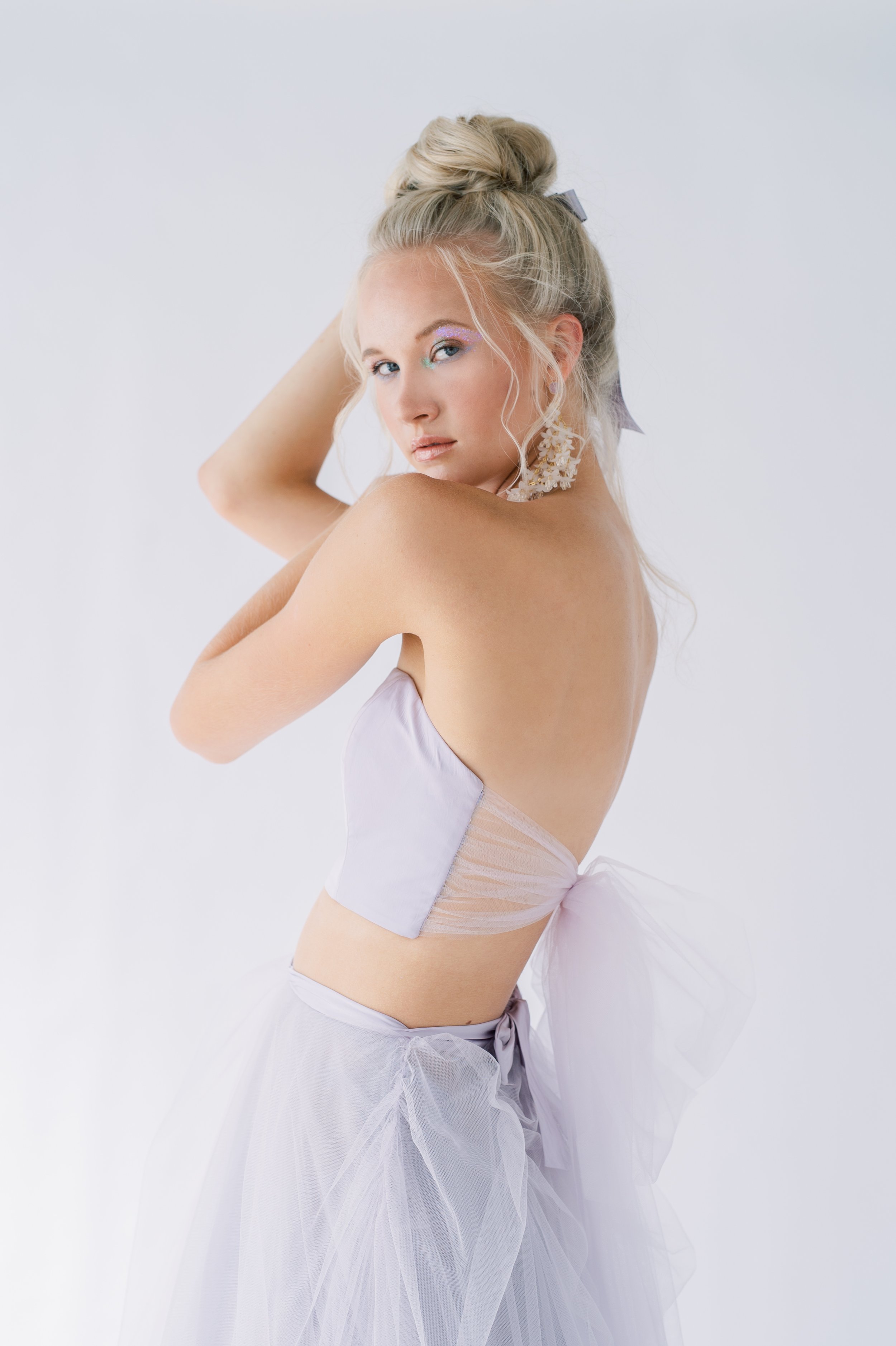 blonde model in a pale lilac bandeau top and lilac tulle overskirt, wearing long dangly white flower earings and a bun in her hear- with a lilac and shimmery eye makeup and glossy lips.