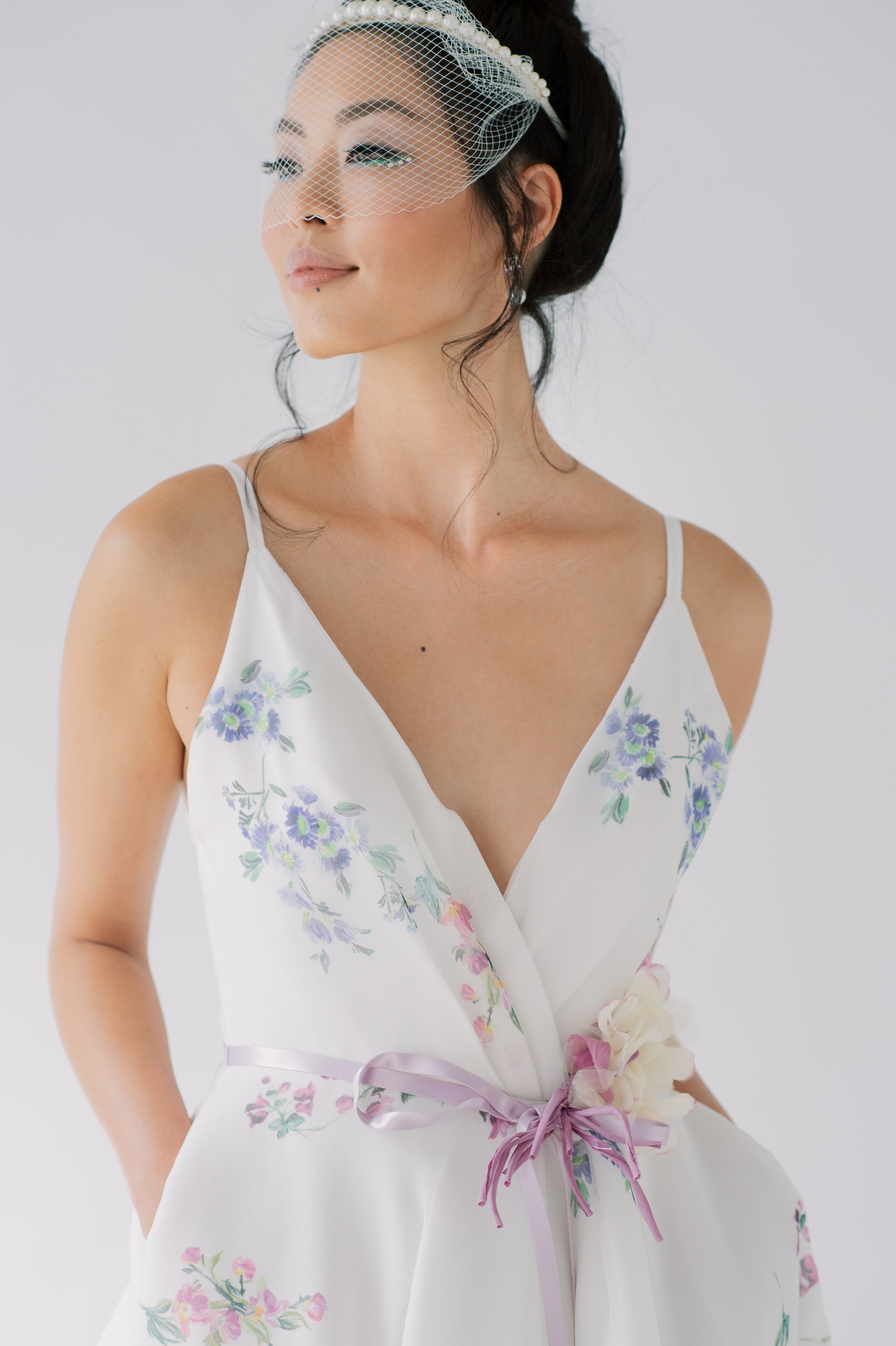 model with black hair up in a bun, with loose pieces falling out, wearing a bircage veil over a pearl headband that is worn as a visor over her eyebrows, in a  white, v neck gown, with a flower print over it and a velvet pink bow at the waistline.