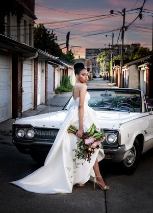 bride stands in alleyway in front of a white vintage car, holding a bouquet