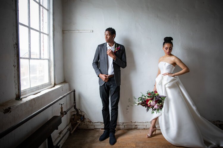 Beautiful bride and groom stand by the window. Groom looking outside, bride looking down. She is wearing a white dress with a wide train and tight bodice and a big bow at the waist, holding a large bouquet of flowers in red and pink tones.