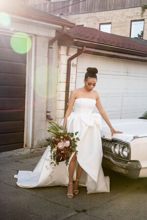 bride looks down at her sandals, in her wedding dress, holding bouquet in the sunset. leaning against vintage white car.